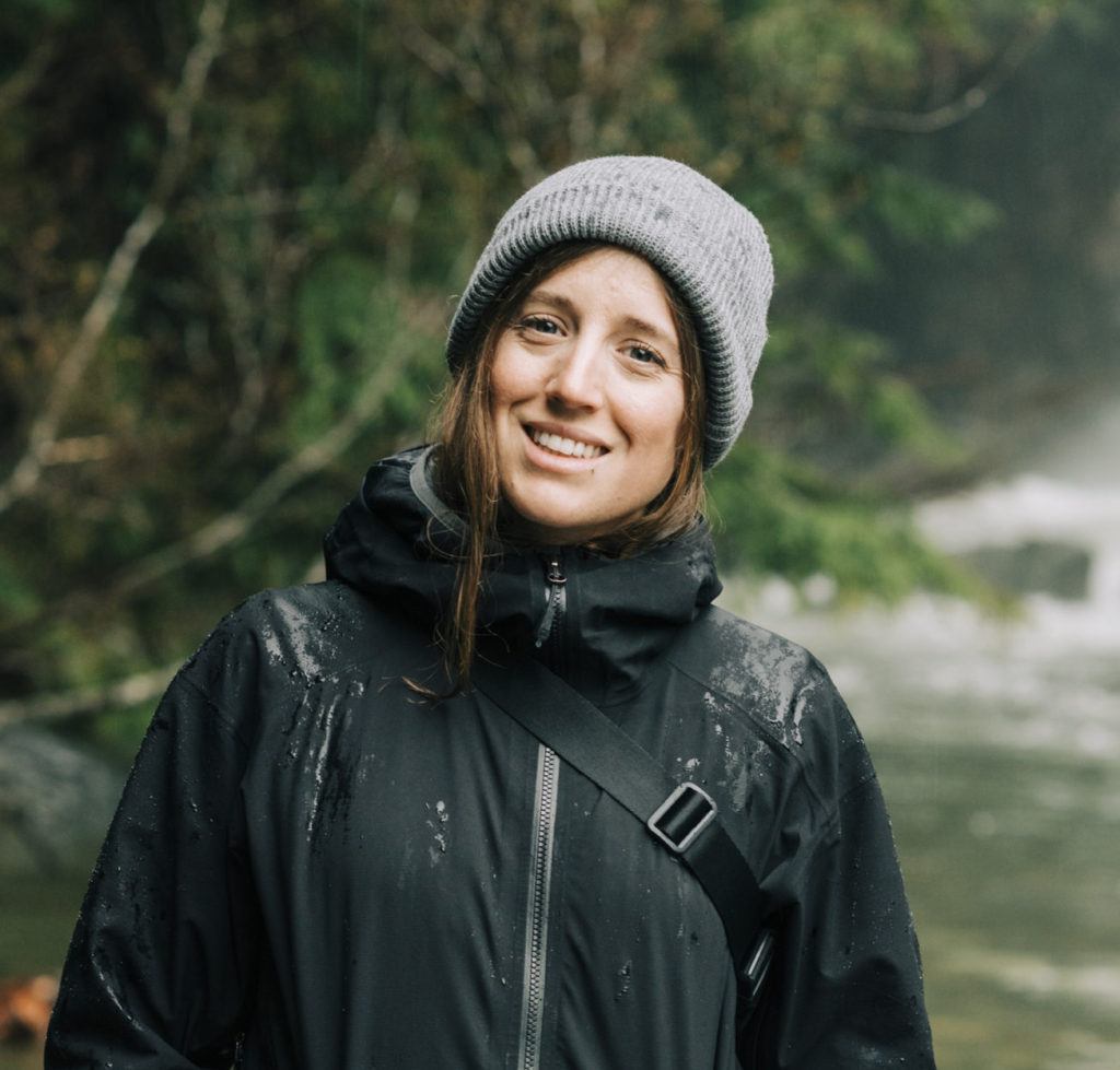 ID: Kate smiles on rainy forest trail wearing grey knit hat and black jacket.