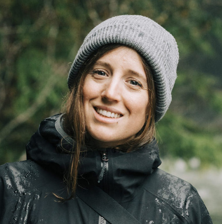 ID: Kate smiles on rainy forest trail wearing grey knit hat and black jacket.