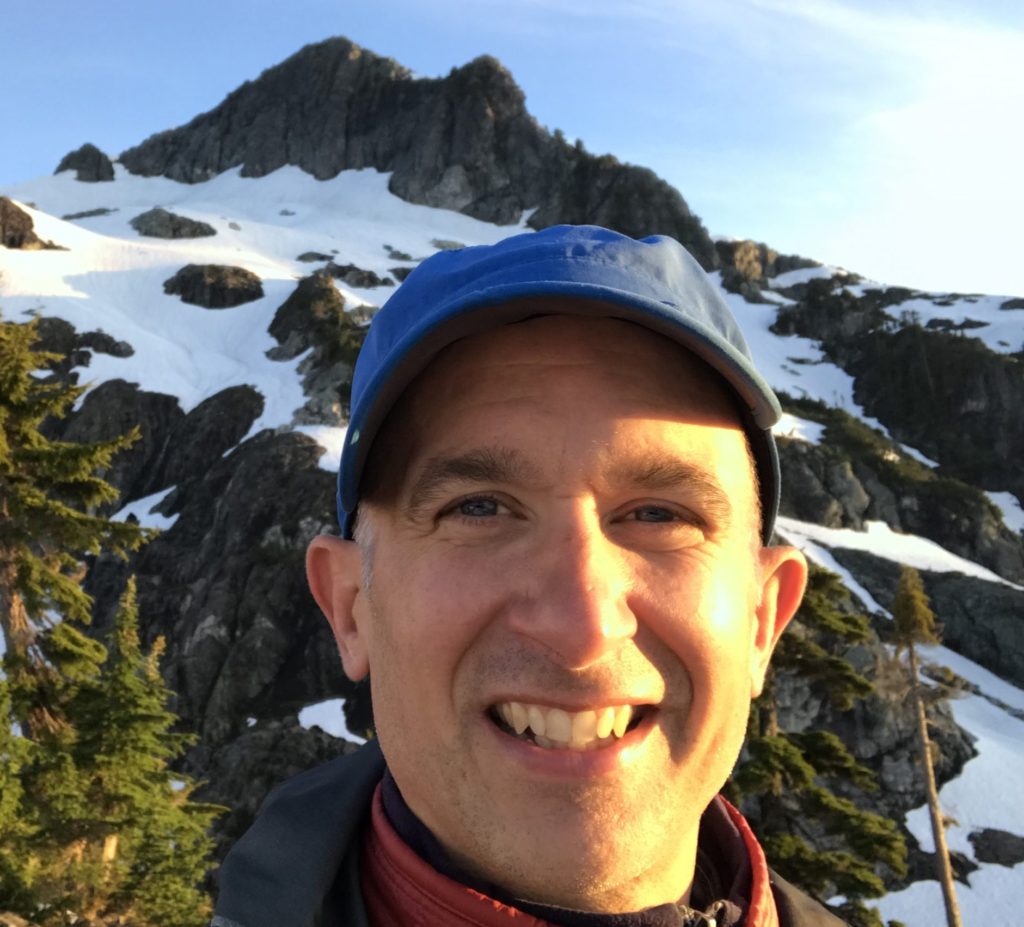 ID: Bruce smiles at Golden Ears mountaintop wearing blue hat.