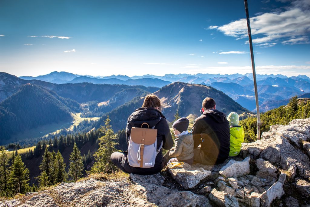 ID: Two adults and two young children sit on mountain top overlooking sunlit mountain valley.