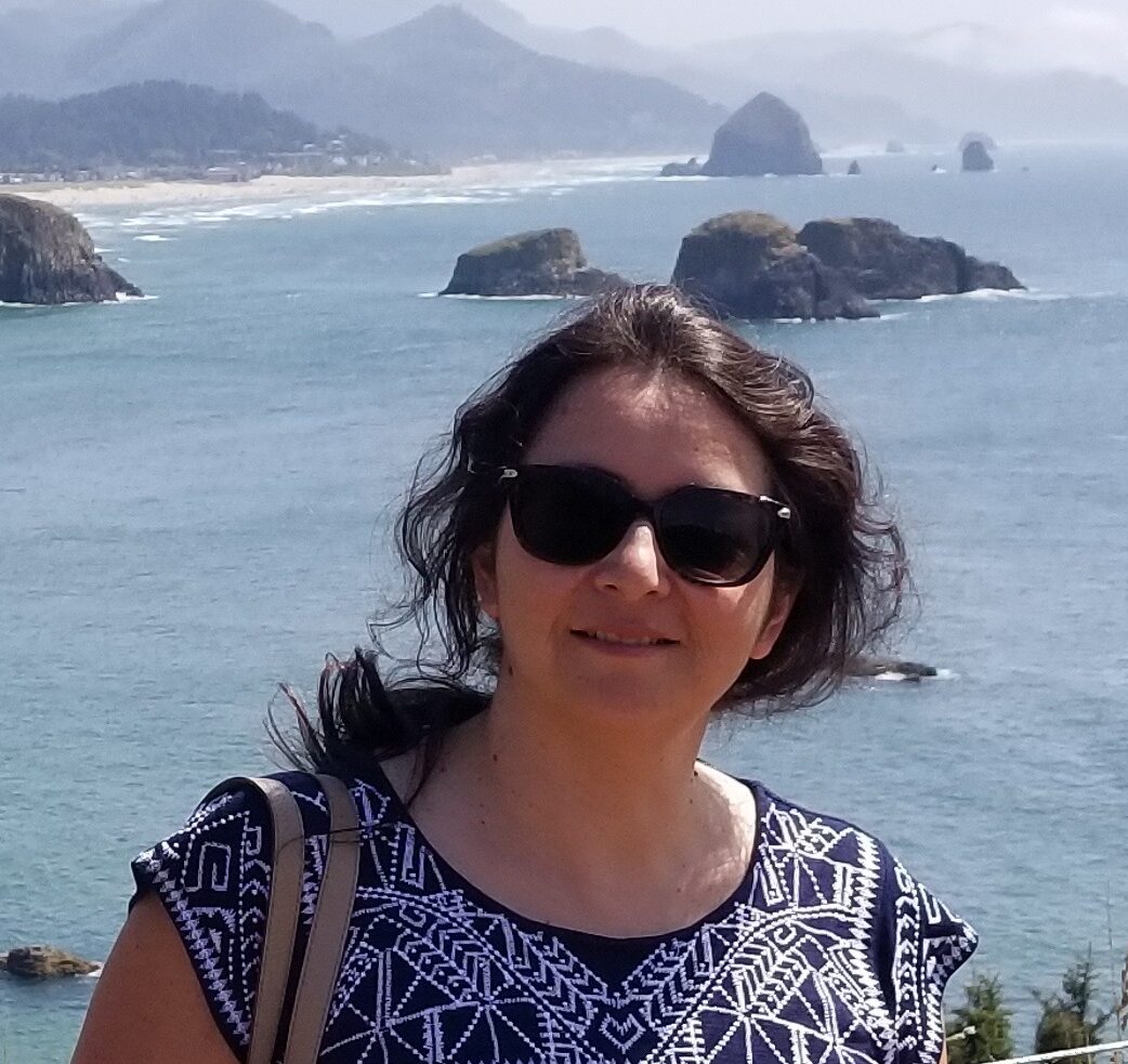 ID: Carla smiles over coastal cliffs wearing dark sunglasses because it's sunny!