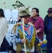 ID: Floyd stands in regalia including blue and red beaded top and headdress. He holds a wooden staff.