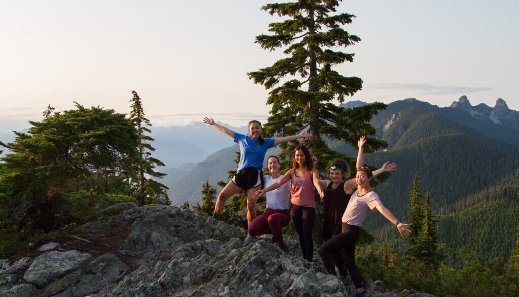 Five f20-somethings friends sit and stand with smiles and airms raised over mountain sunset.