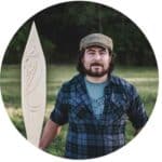 ID: Jessie wear blue button shirt and brown cap. Holding carved wood paddle by forested backdrop.