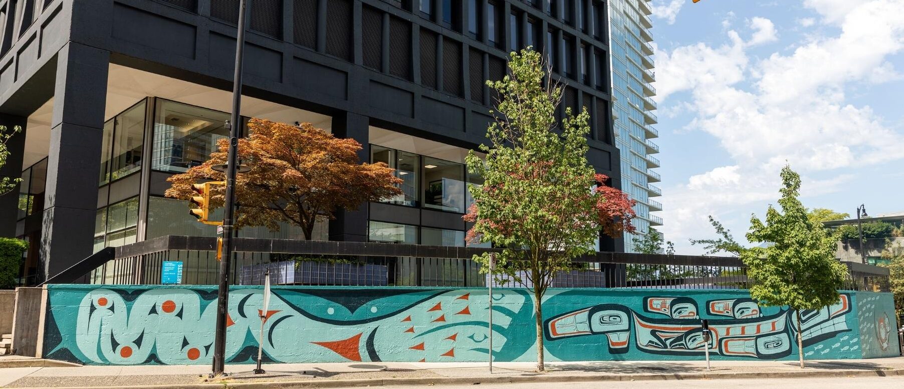 ID: Shades of blue and green if form line art features whale, otter and euchalon fishes on long sidewalk wall.