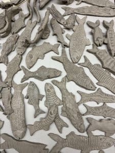 ID: patchwork of unique grey clay fish shapes with scraped tiles and fins lay on a grey table.